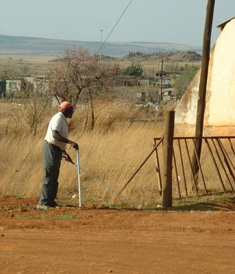 In this photo, the man with the white support cane and the long white cane with a bundu basher tip has reached an opening in the fence and we can see part of a building or large tent on the other side of the fence.